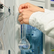 Zwei Hände halten eine Phiole vor einem Laborgerät